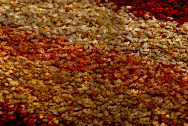 Kolorowy mięcisty dywan w odcieniach brązu i czerwieni z widocznymi moznymi zabrudzeniami.